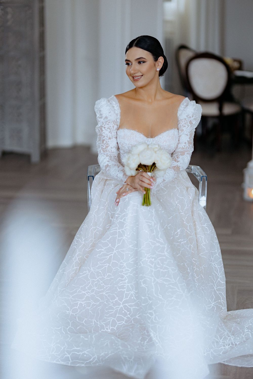 Elegant bride in a white lace ball gown with puff sleeves holds a bouquet in a bright interior setting.