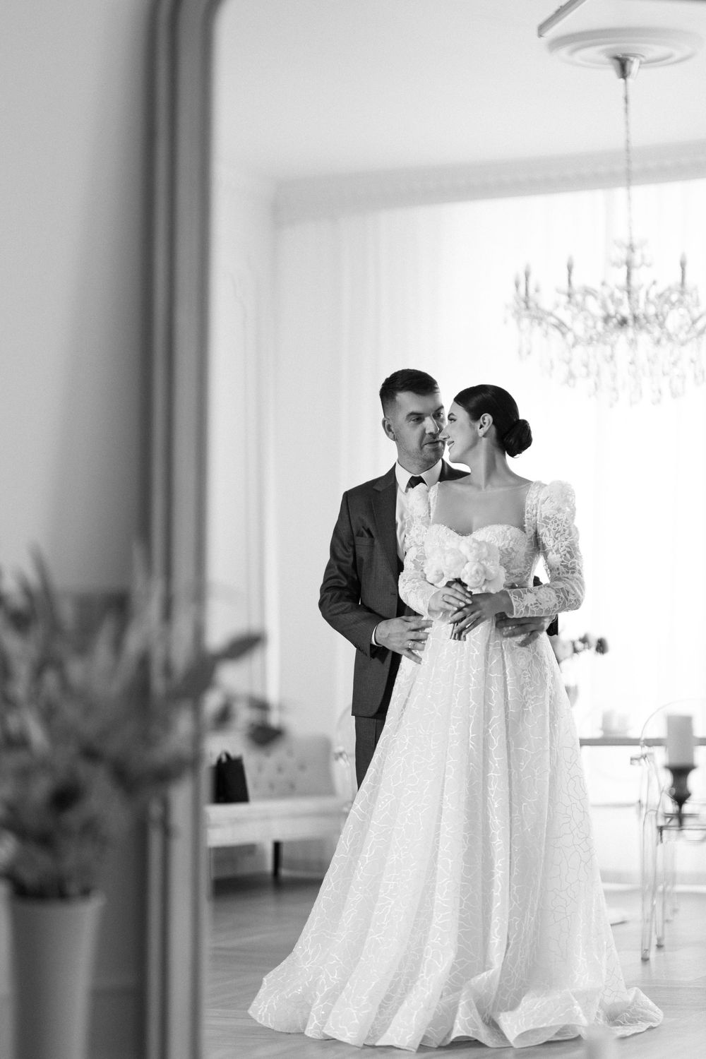 A couple shares a tender dance moment in a bright white ballroom with elegant chandeliers.
