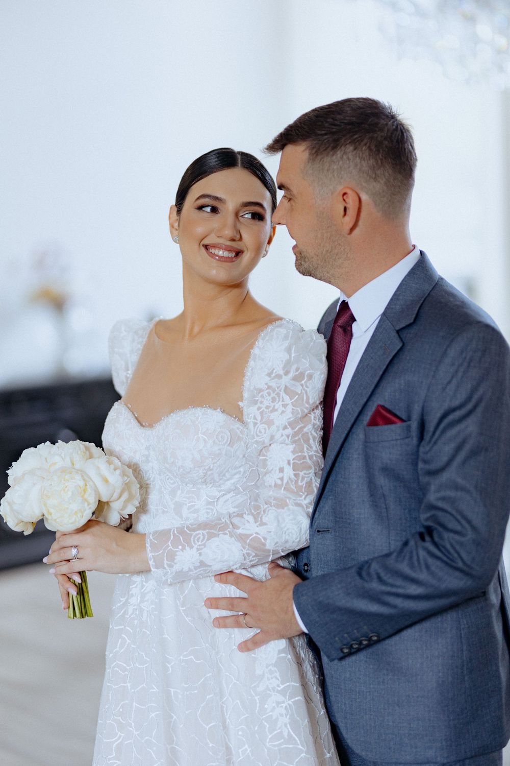 A romantic wedding moment with a couple in white lace gown and grey suit holding a white bouquet.