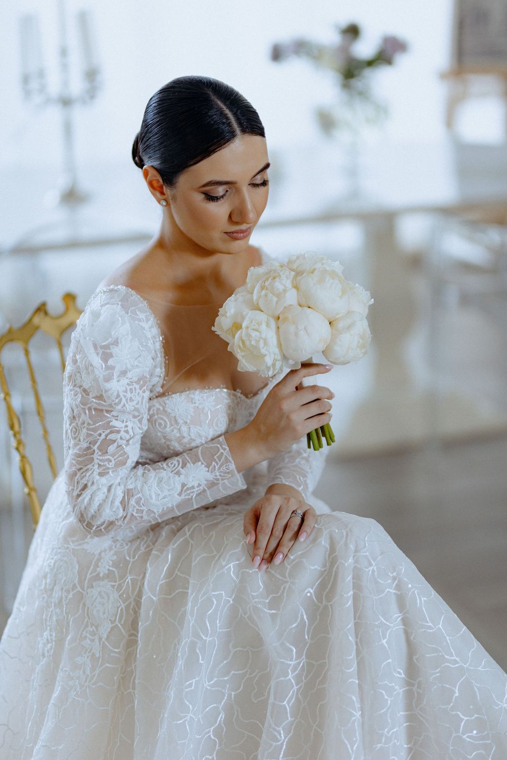 Bride in long-sleeve lace gown sits elegantly with white peony bouquet in bright interior.