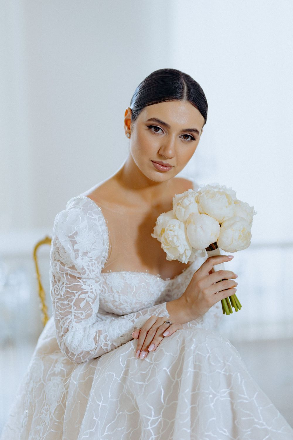 A bride in a white lace long-sleeved wedding gown poses elegantly with a white floral bouquet in a bright setting.