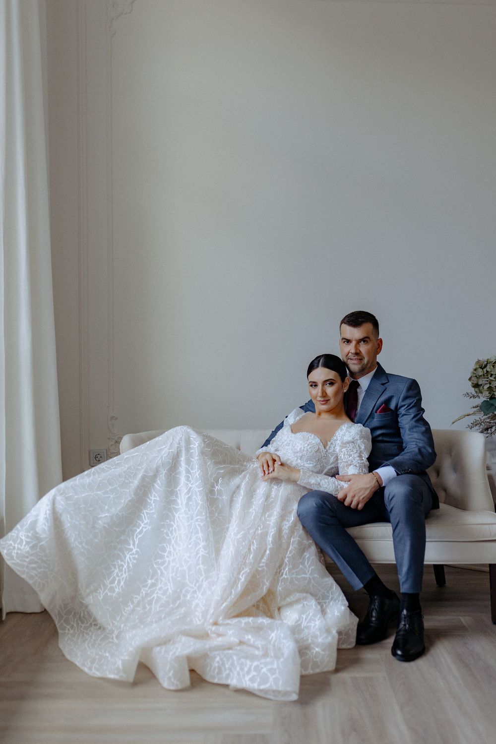 A bride in a white lace gown and groom in navy suit pose artistically on a white couch in a minimalist studio setting.