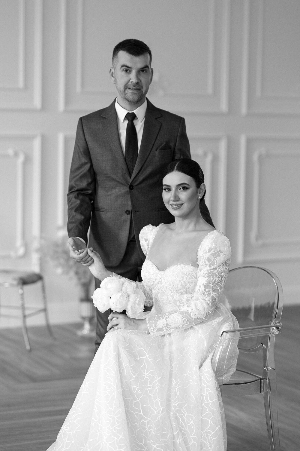 A black and white wedding portrait of a couple in formal attire, with the bride seated in a lace gown holding flowers.