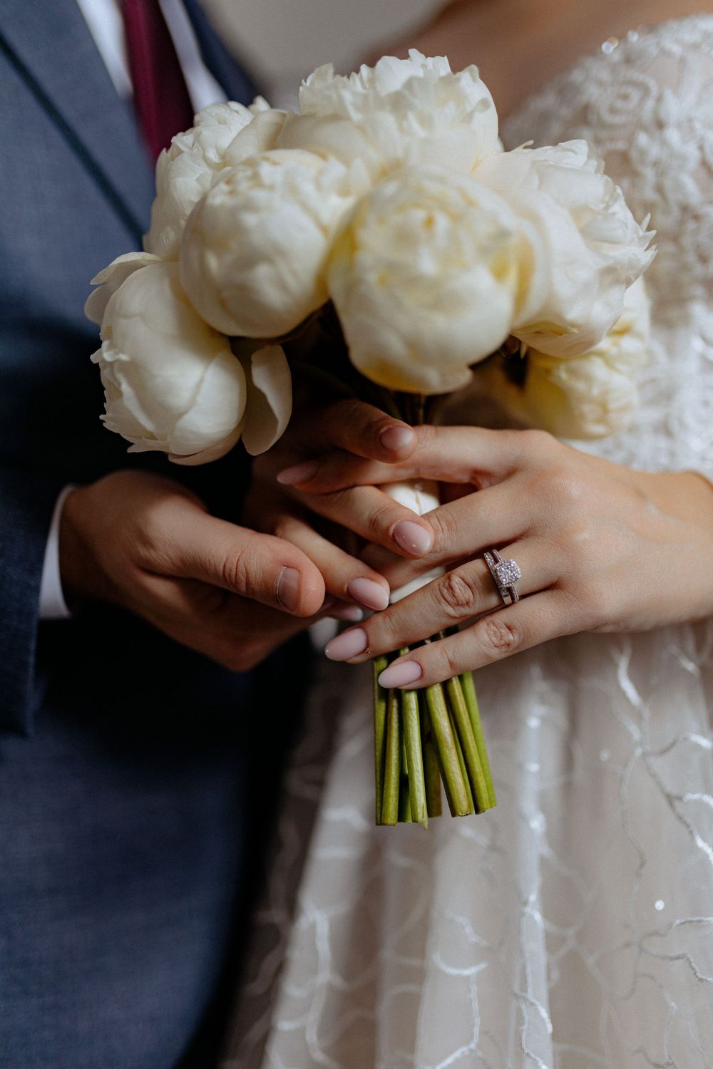 Close-up of white peony bouquet and wedding rings held by bride and groom's hands.