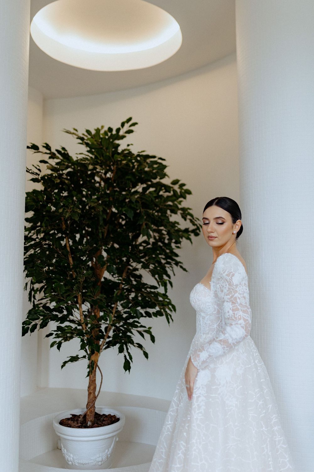 A bride in a luxurious white lace ball gown poses elegantly against white columns and potted plants.