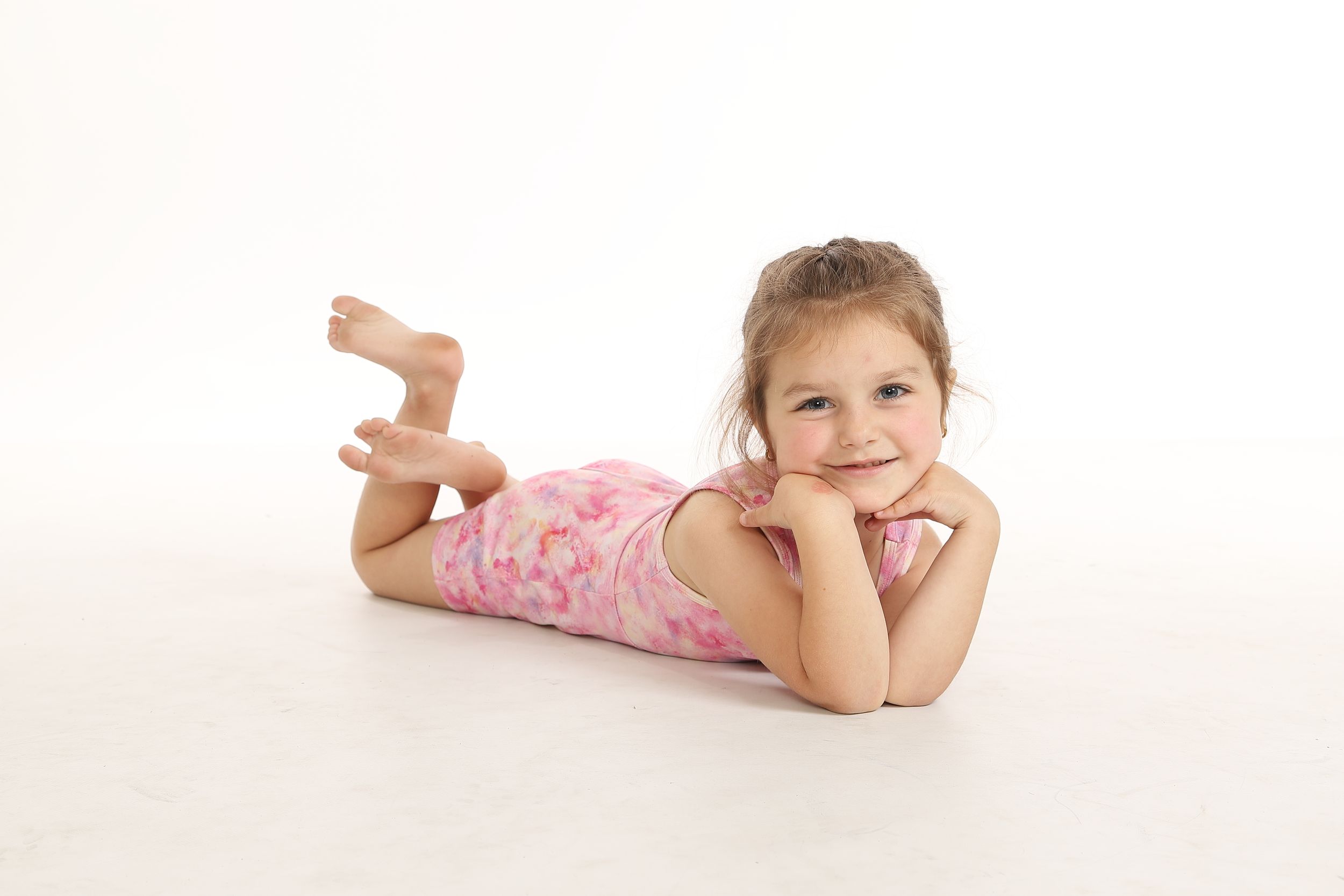 A child in a pink polka dot dress lies on their stomach on a white floor, smiling at the camera.