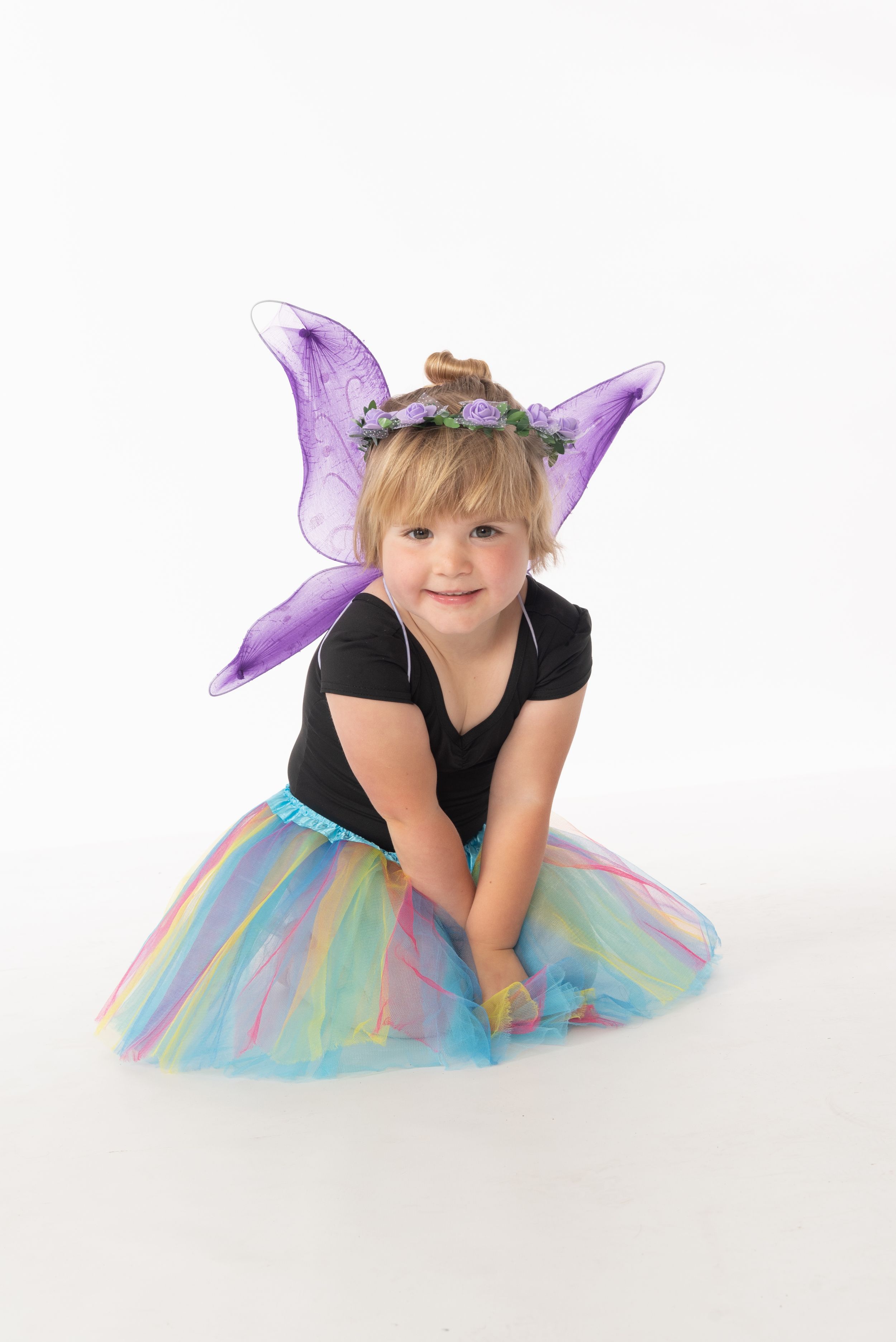 A dancer in a black leotard and pastel tulle tutu with purple fairy wings poses against a white backdrop.