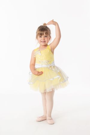 Young dancer in yellow tutu dress and pink ballet shoes poses gracefully against white background.