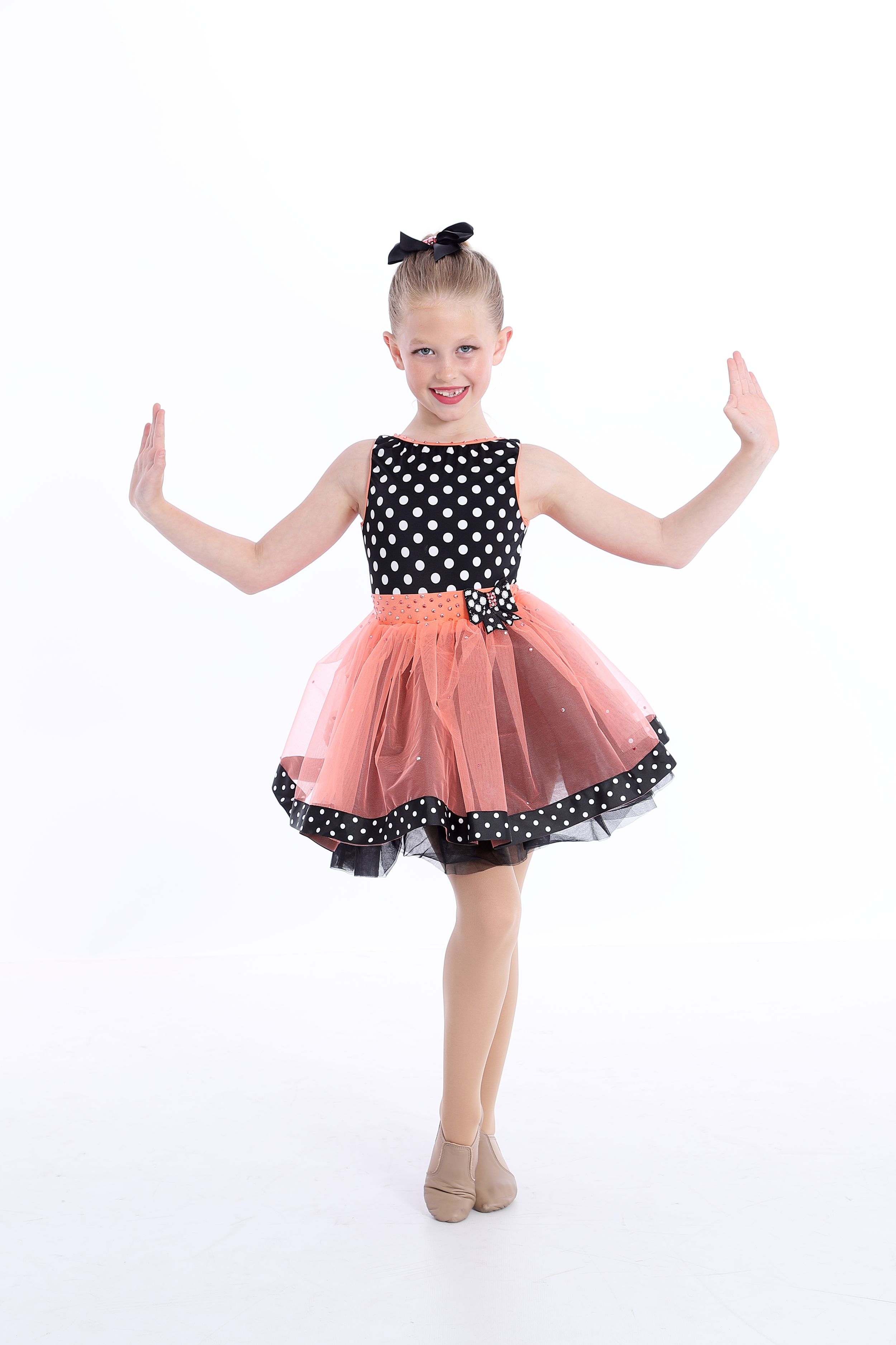 Young dancer in polka dot and peach tulle dress strikes a joyful pose against white background.