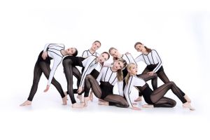 Group of dancers in black and white outfits pose together in modern contemporary dance formation against white backdrop.