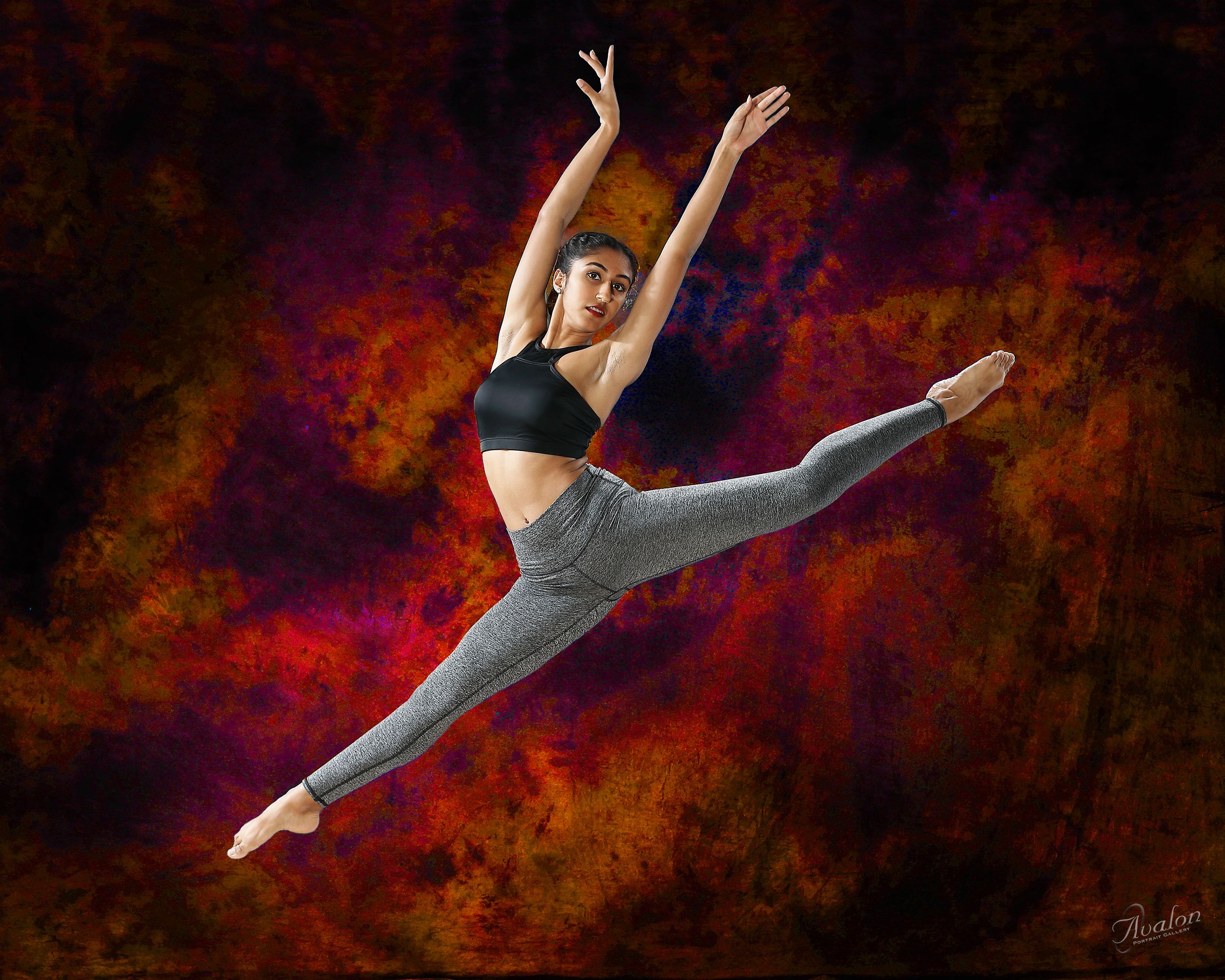 A ballet dancer in grey leggings and black top leaps gracefully through the air against a dark red and orange background.
