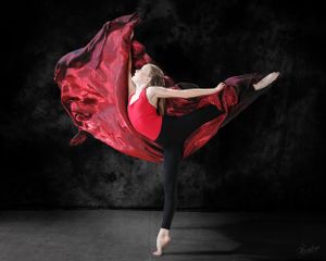 A ballet dancer in red costume performs an elegant arabesque with flowing fabric against a dark background.