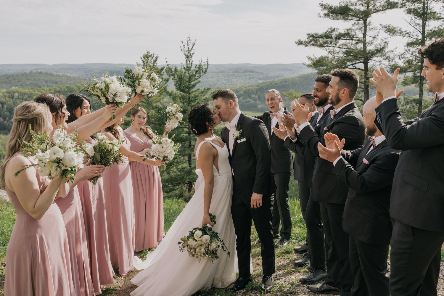 Wedding party in pink dresses and black suits celebrates on hilltop with scenic mountain view.