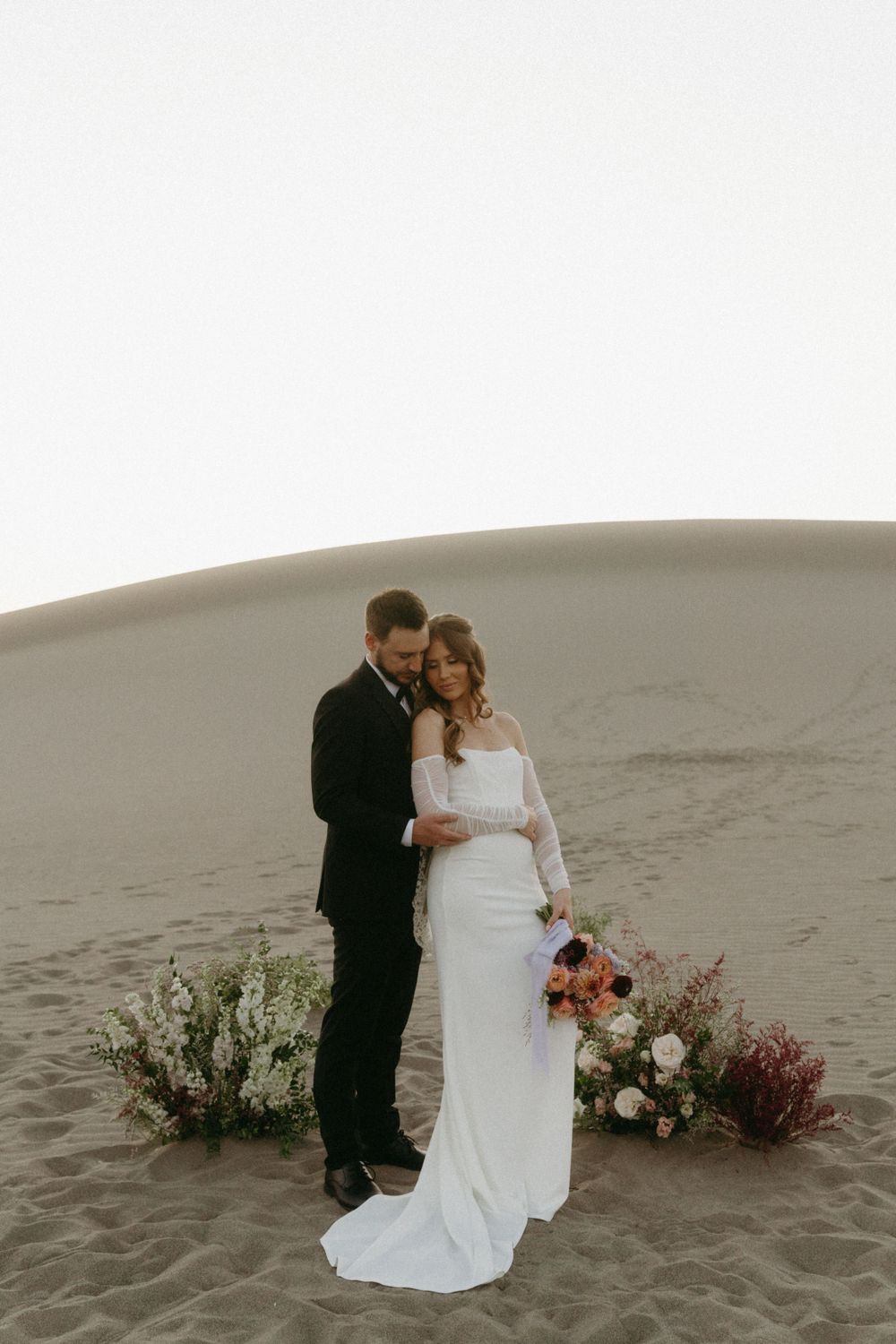 A wedding couple embraces on sand dunes with floral arrangements at their feet.