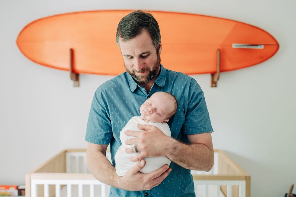 A dad holds a sleeping newborn baby against a white background with an orange surfboard mounted on the wall during Orlando newborn session.
