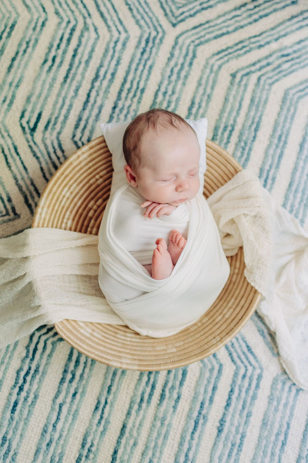 A sleeping newborn baby wrapped in white fabric rests peacefully on a woven basket against a blue striped background.