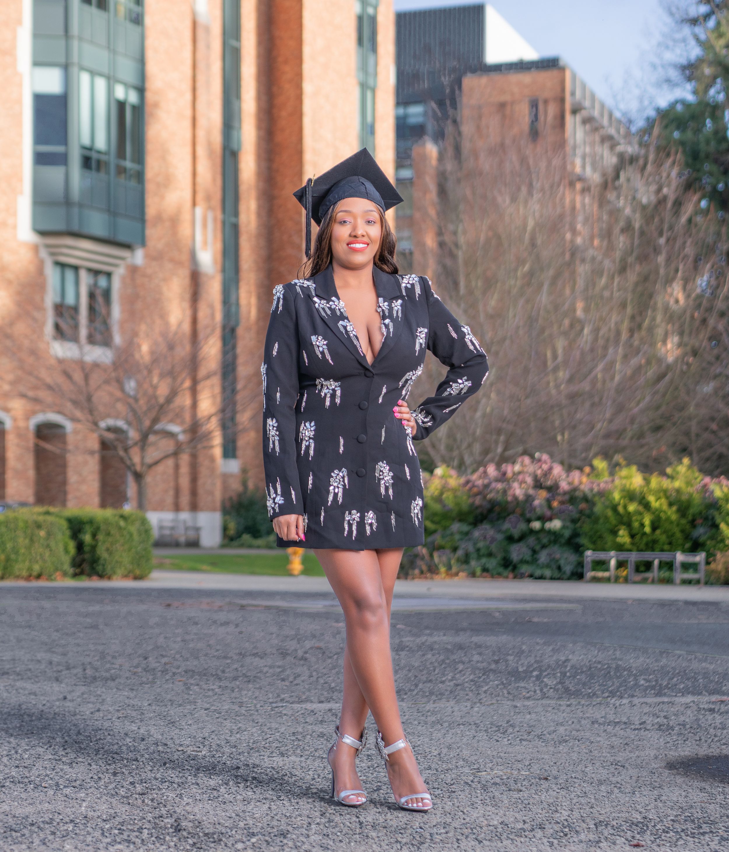 A person in a black dress and boots poses playfully on a paved walkway near brick buildings.