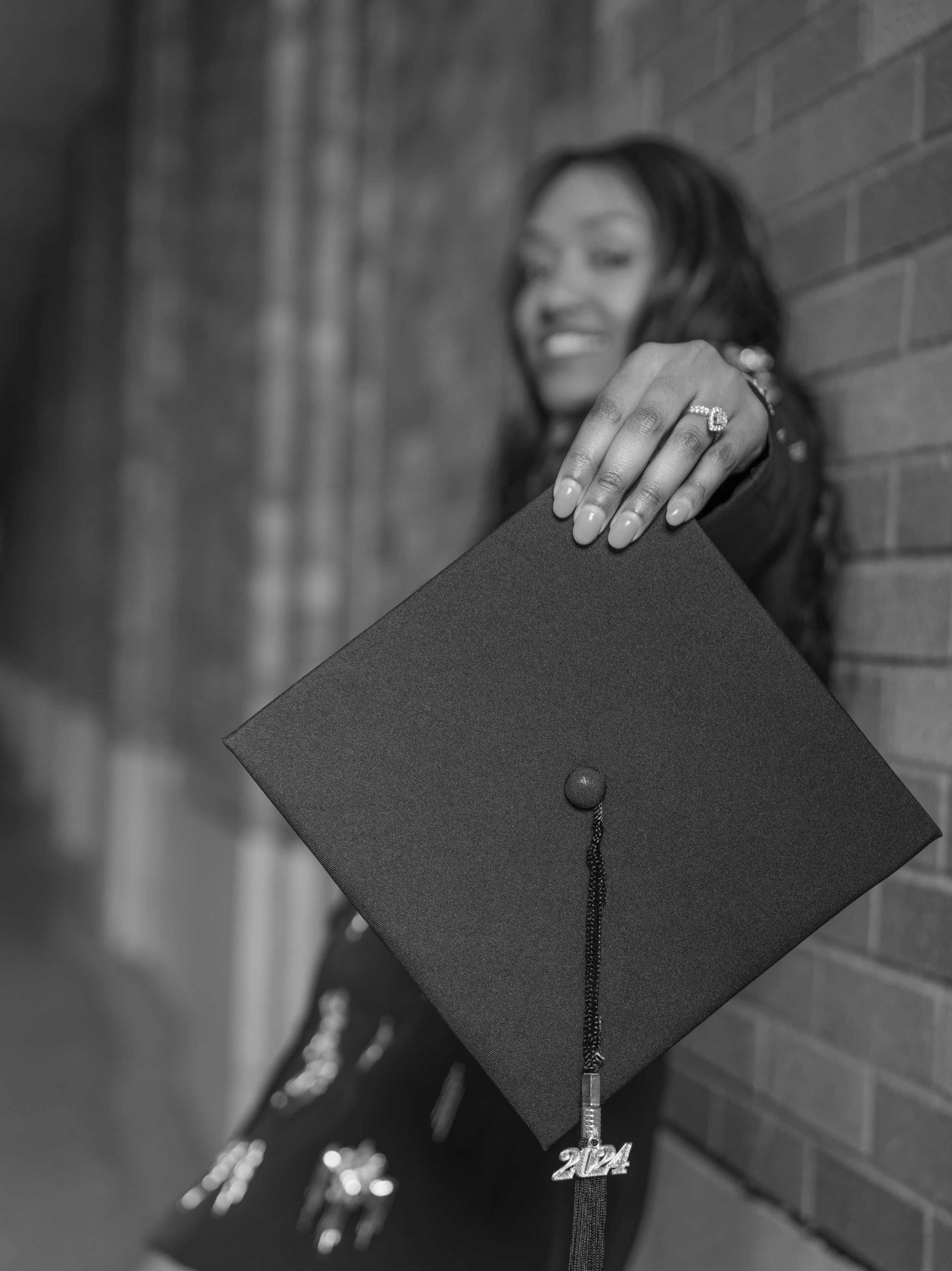 A graduation cap with tassel is held up against a brick wall in an artistic black and white photo.