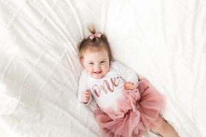 A baby wearing a pink tulle dress lies on white bedding while holding a pink cotton candy heart.