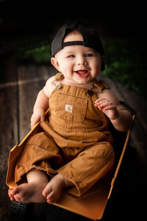 A joyful baby wearing brown Carhartt overalls and a backwards baseball cap sits on a wooden surface outdoors.