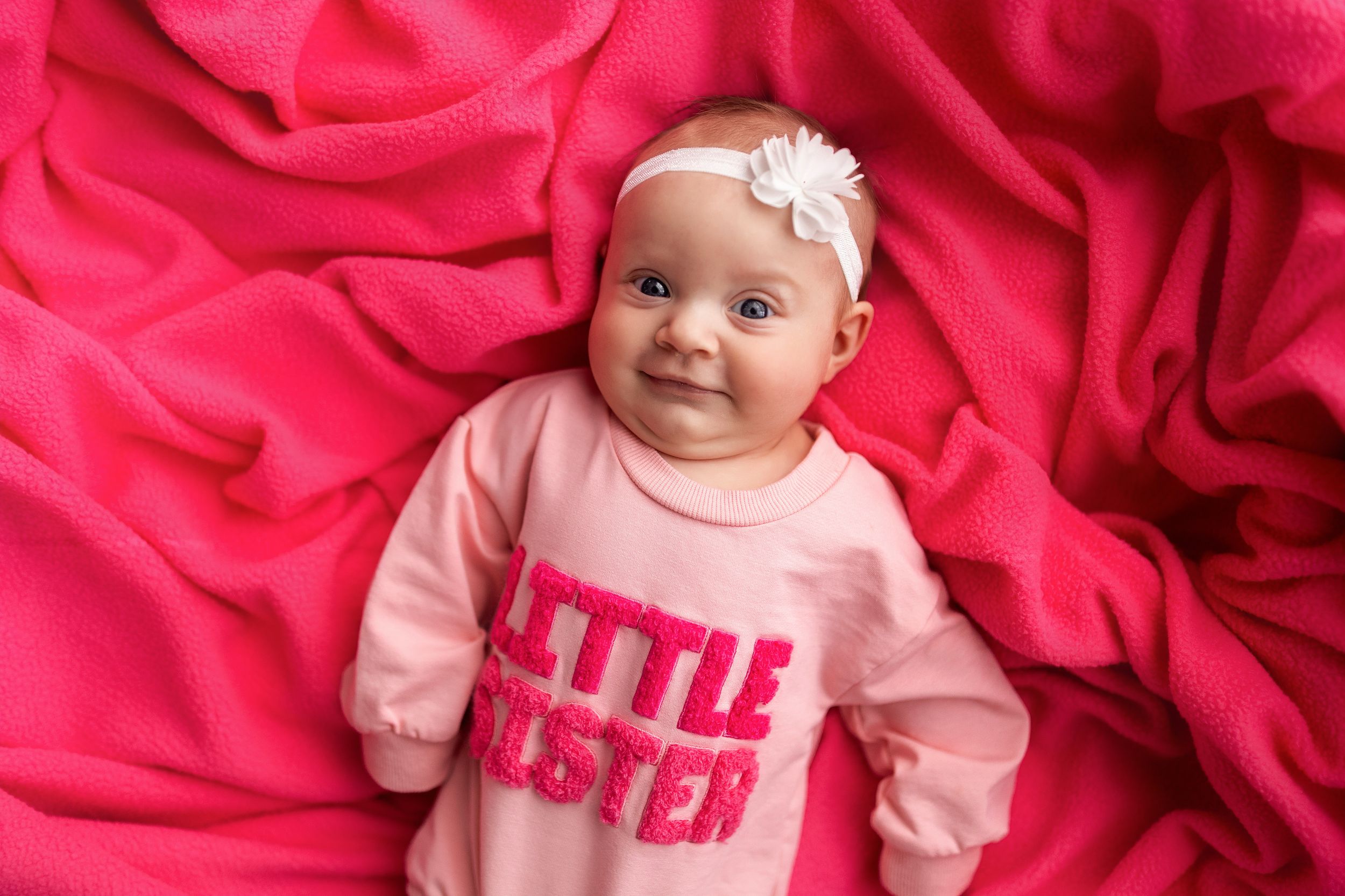 A baby wearing a pink outfit lies on bright pink fabric while looking at the camera.