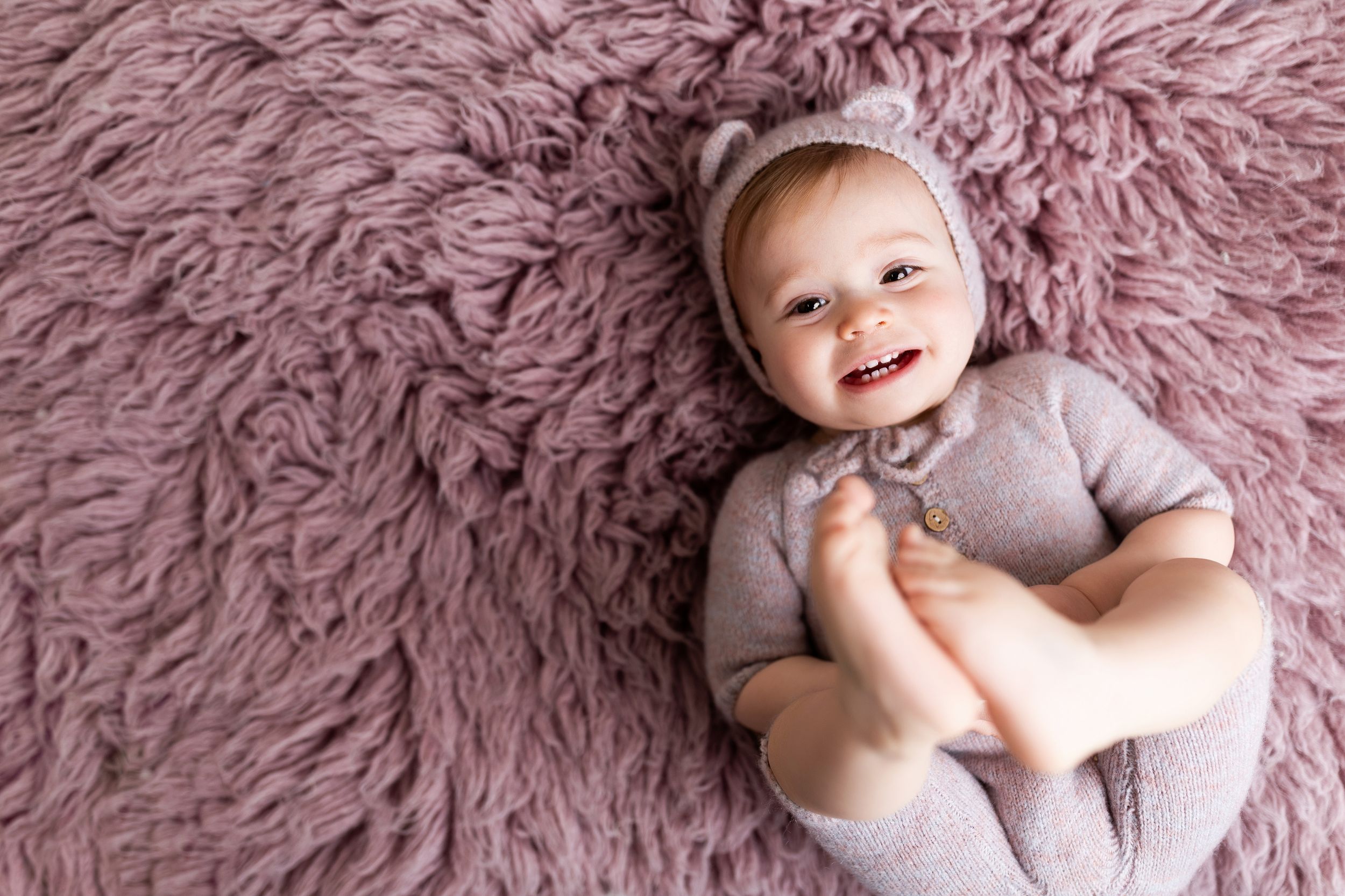 A happy baby wearing a gray onesie lies on a plush mauve-colored furry rug while smiling at the camera.
