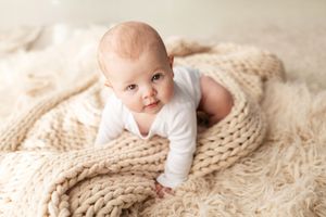A baby wearing white lies on a cozy cream knit blanket in a bright studio setting.