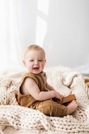 Baby wearing brown outfit lying on chunky knit cream blanket while smiling and playing.