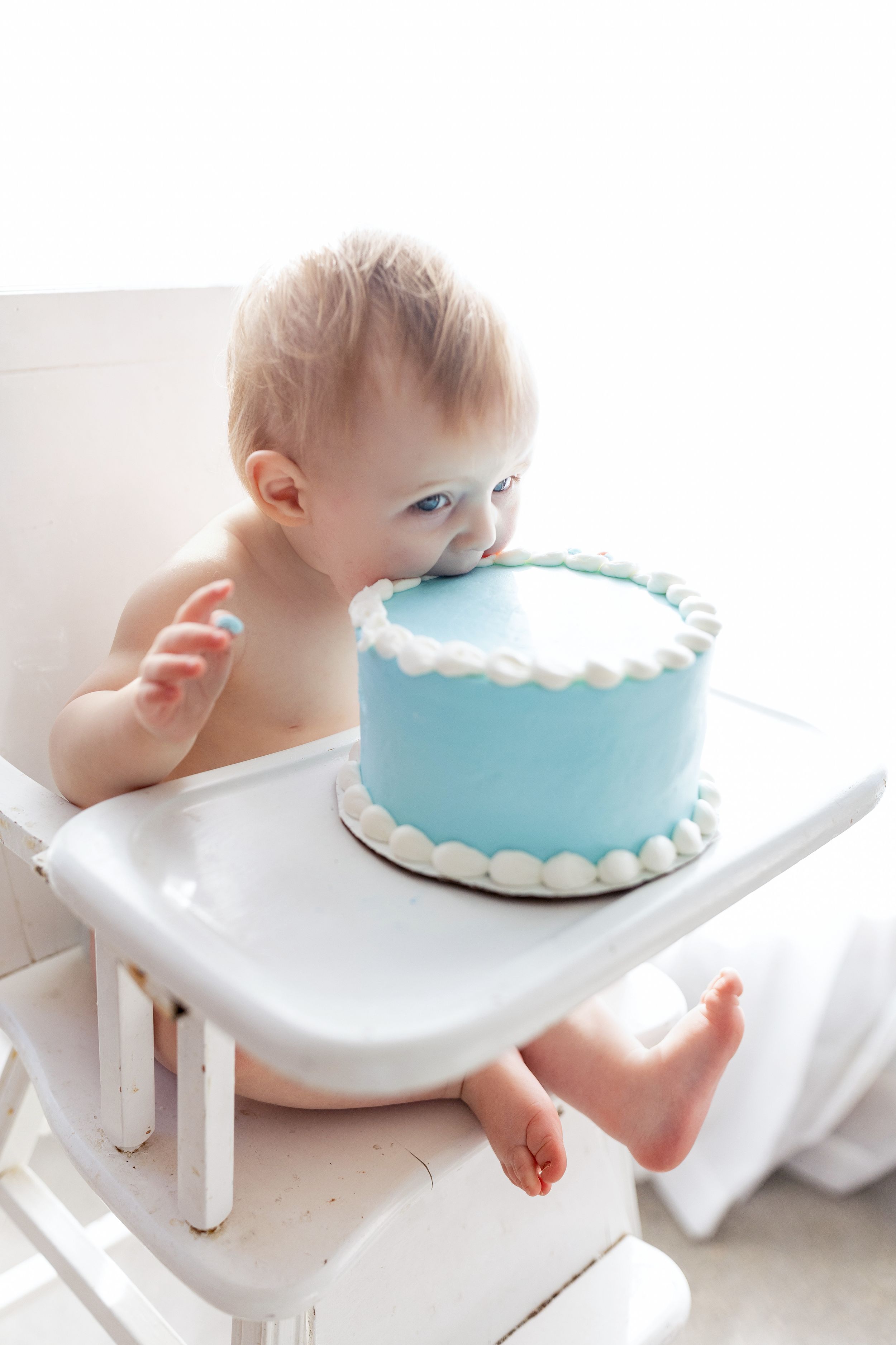 Baby in white high chair leaning towards light blue birthday cake with white scalloped frosting decoration.