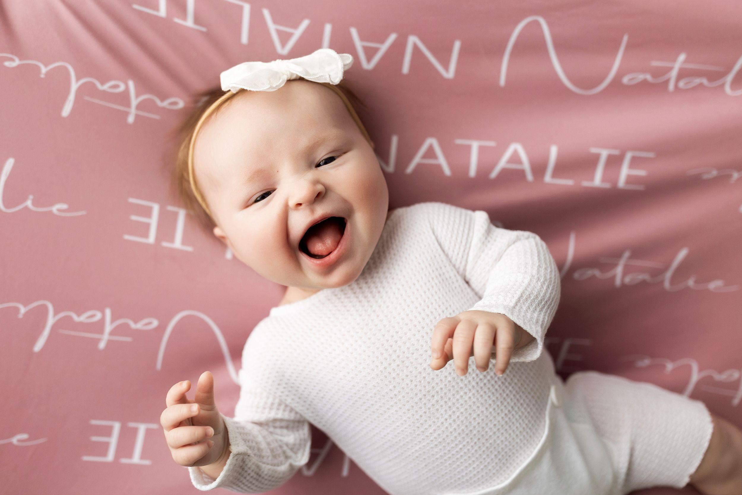 A laughing baby wearing white lies on a pink background with text overlay.