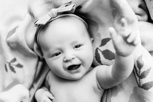 A joyful baby wearing a white bow headband lies on patterned fabric while smiling and reaching up.