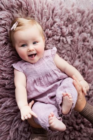 A baby in a lavender dress lies on a plush purple rug while laughing and playing.