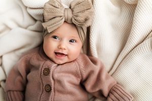 A baby wearing a cozy brown knit sweater lies on soft white textured bedding.