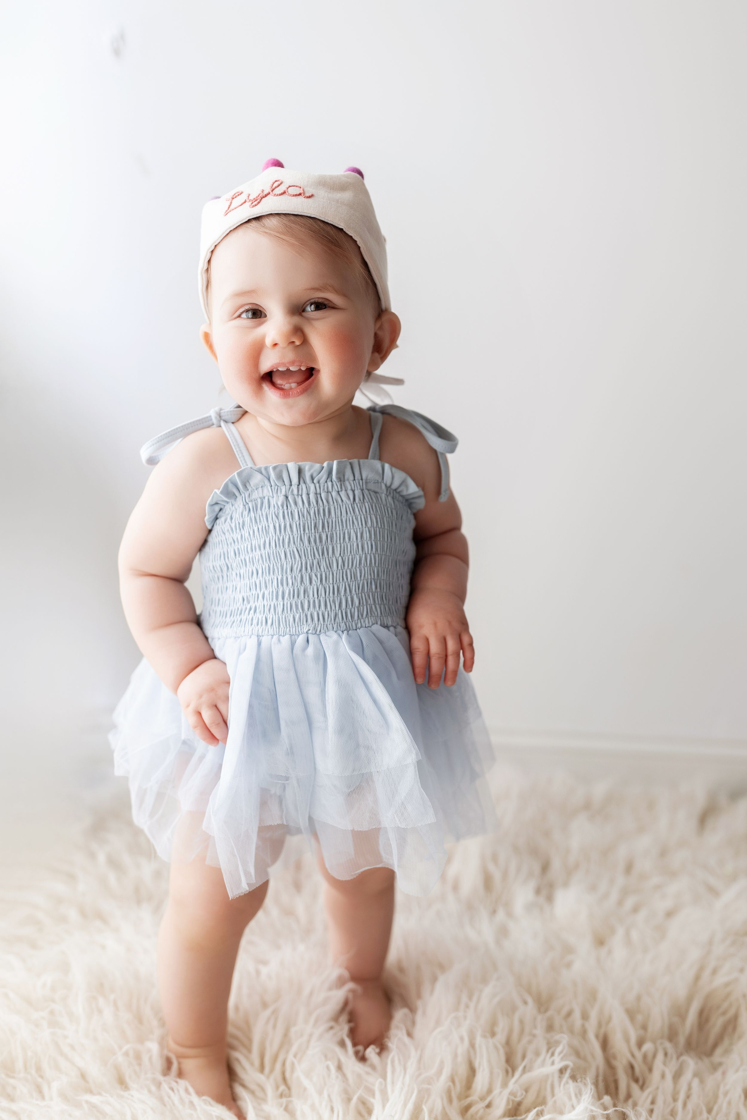 A baby in a light blue and white smocked dress smiles and plays on a fluffy white rug in a series of photos.