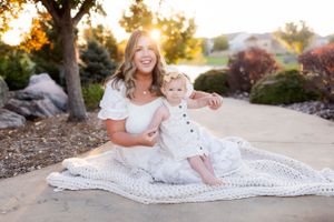 A mother and child sit together on a white blanket during golden hour in a garden setting.