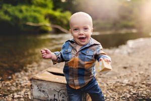 A joyful baby in denim and plaid laughs while playing outdoors by a stream.