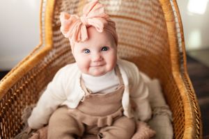 Baby in cream sweater and pink bow sitting in wicker chair.