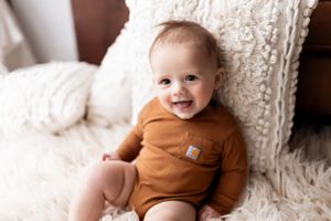 Smiling infant wearing brown onesie sitting against white textured blanket.