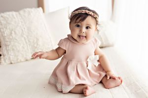 Baby wearing pink dress and headband sits on white couch with decorative pillow.