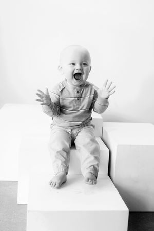 Black and white photo of a joyful infant sitting on a white block with hands raised and mouth open in excitement.