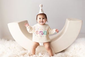 Baby in white 'one' outfit sitting on curved white rocker against white backdrop.