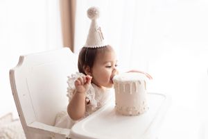 Small child in party hat reaching toward white birthday cake on high chair.