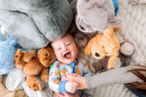 Happy infant lying on a bed surrounded by plush teddy bears and soft toys.