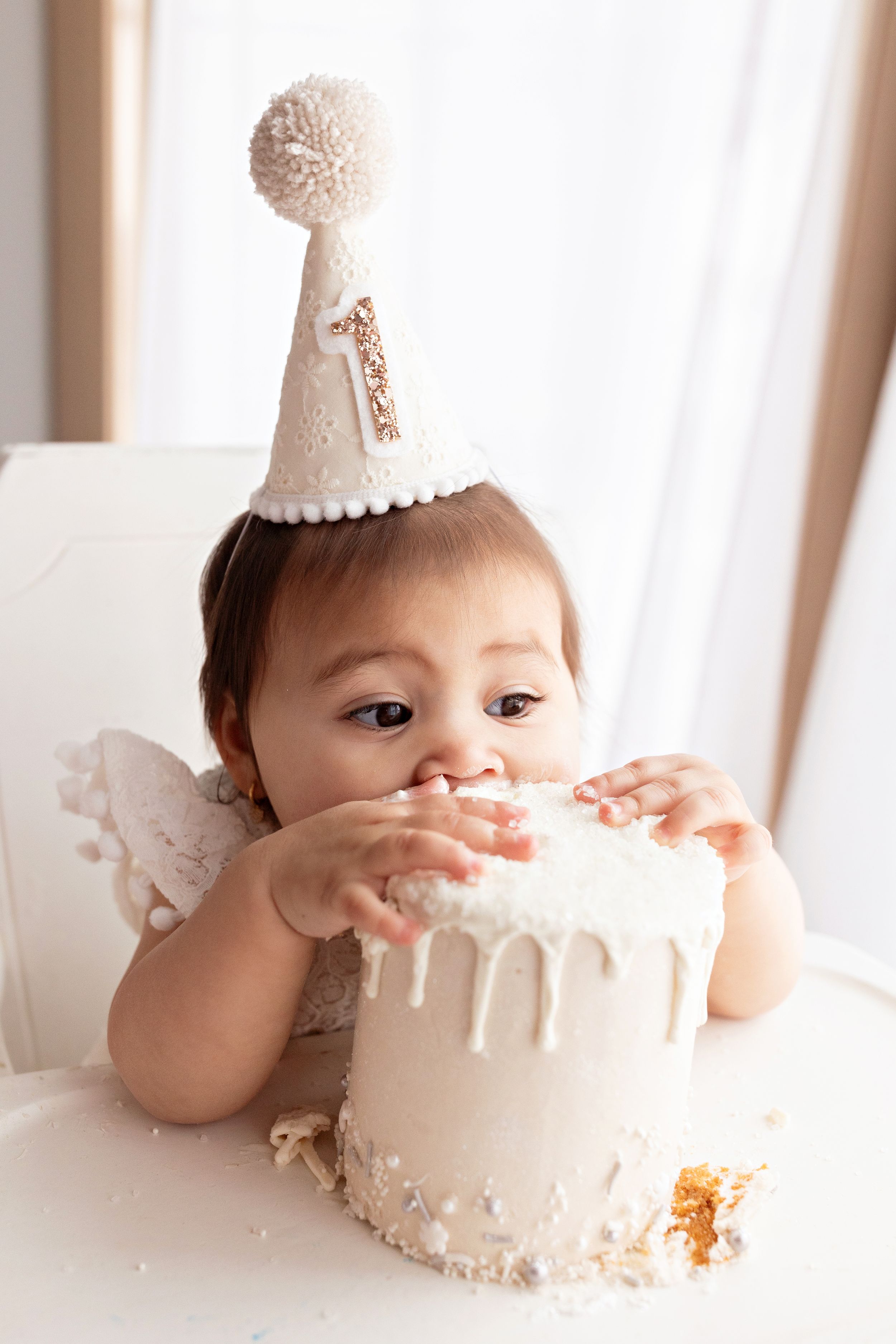 Messy and adorable cake smash photo with white drip cake and party hat.