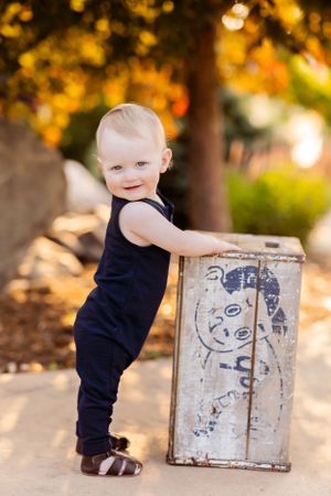 A toddler in navy overalls leans against a rustic box outdoors at sunset.