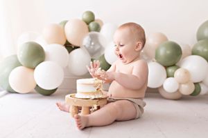 Baby in diaper sits with cake surrounded by pink and green balloons.