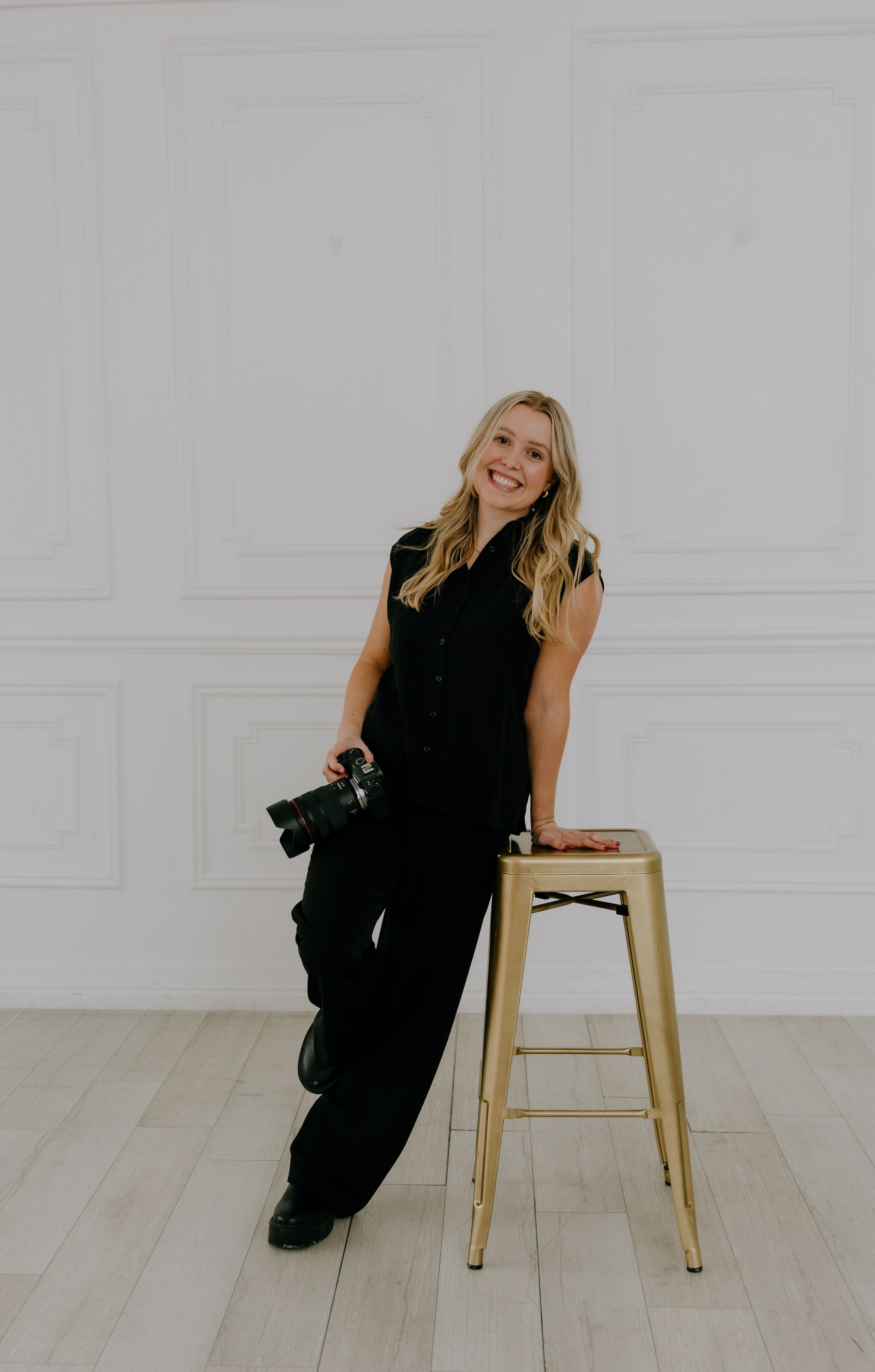 Blonde female photographer in black jumpsuit posing with camera beside gold stool in white studio.