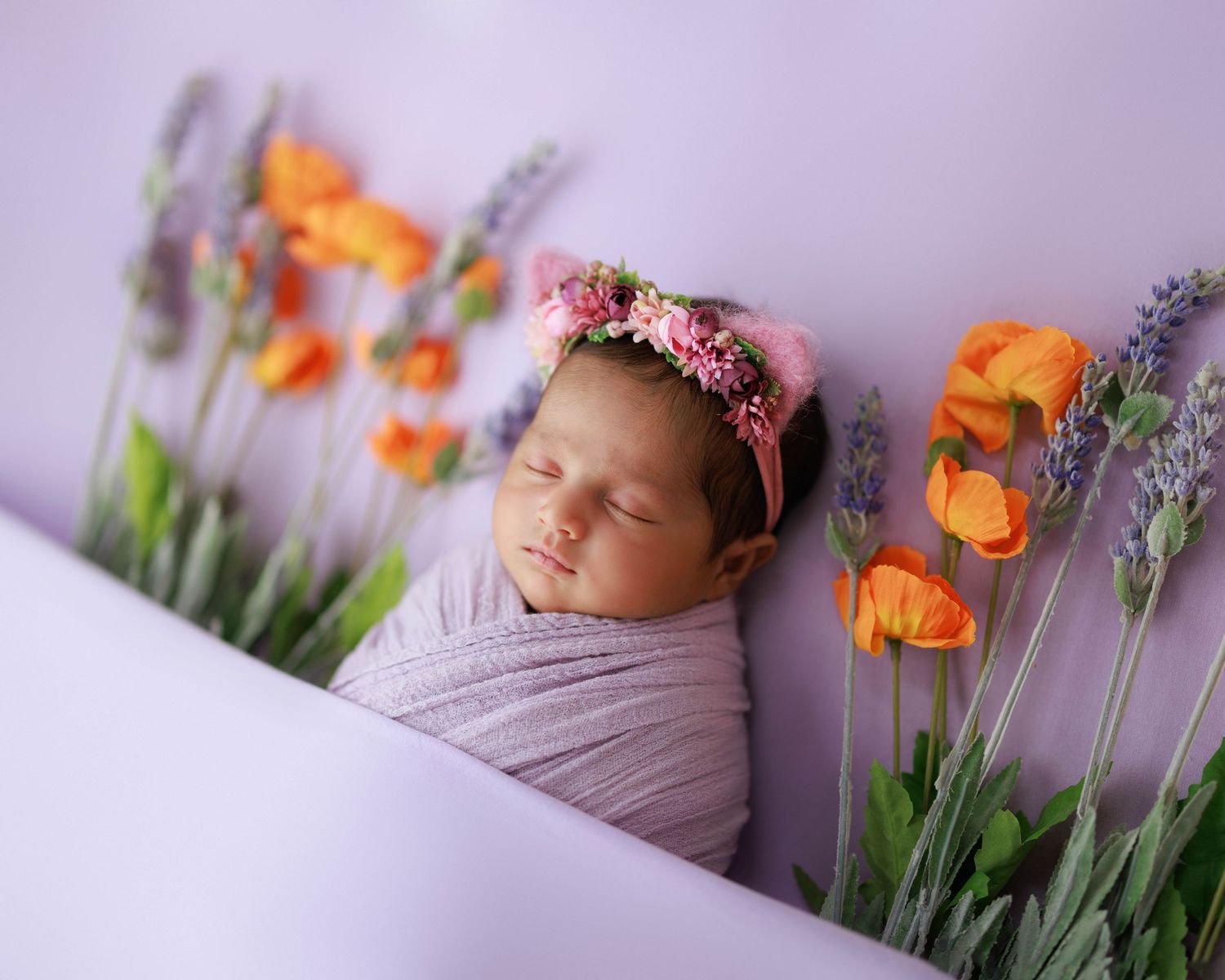 A sleeping newborn wrapped in purple swaddle with orange tulips and lavender flowers as decoration.
