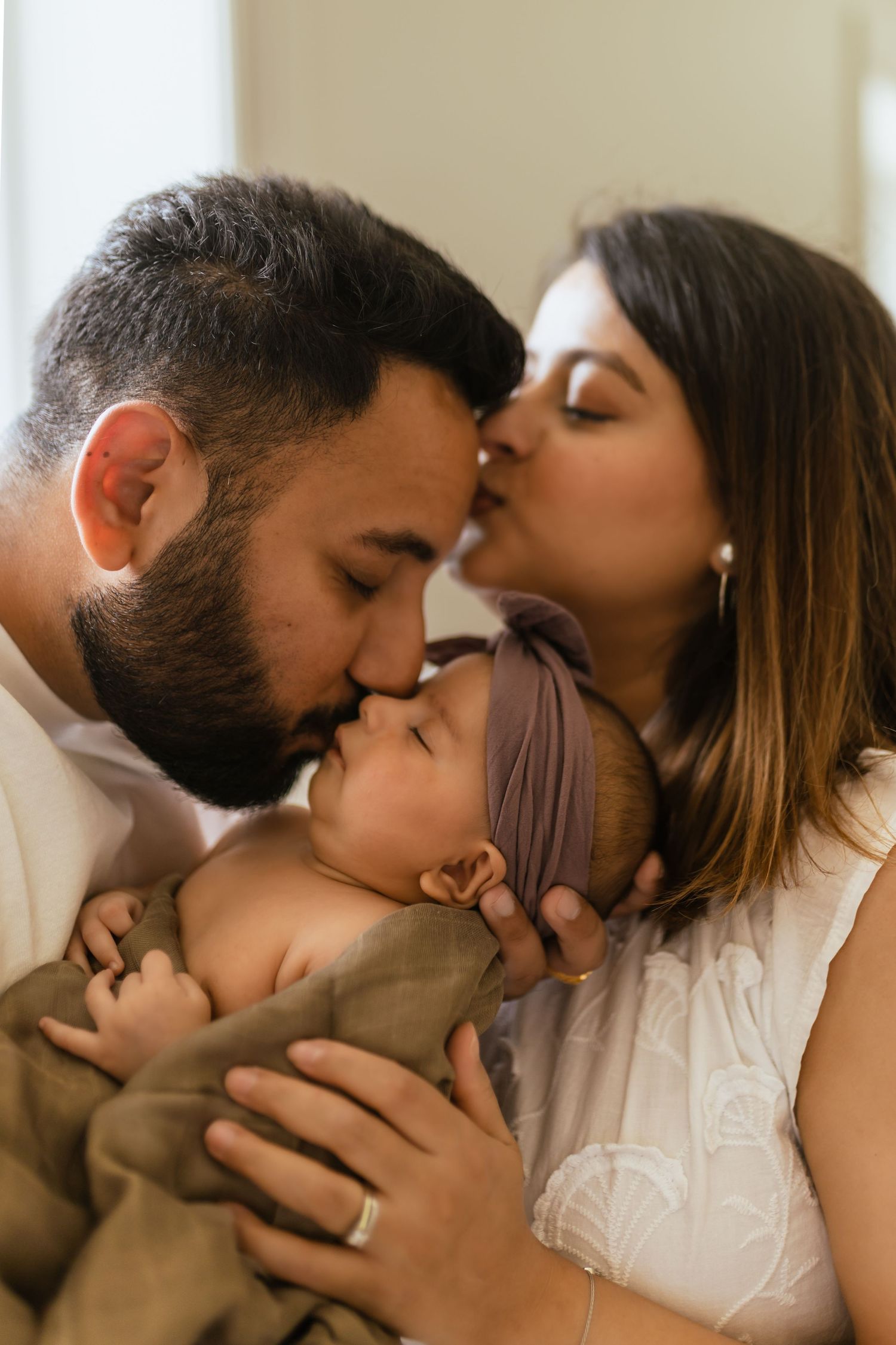 A couple sharing a tender moment while kissing their baby in a warm, intimate family portrait.
