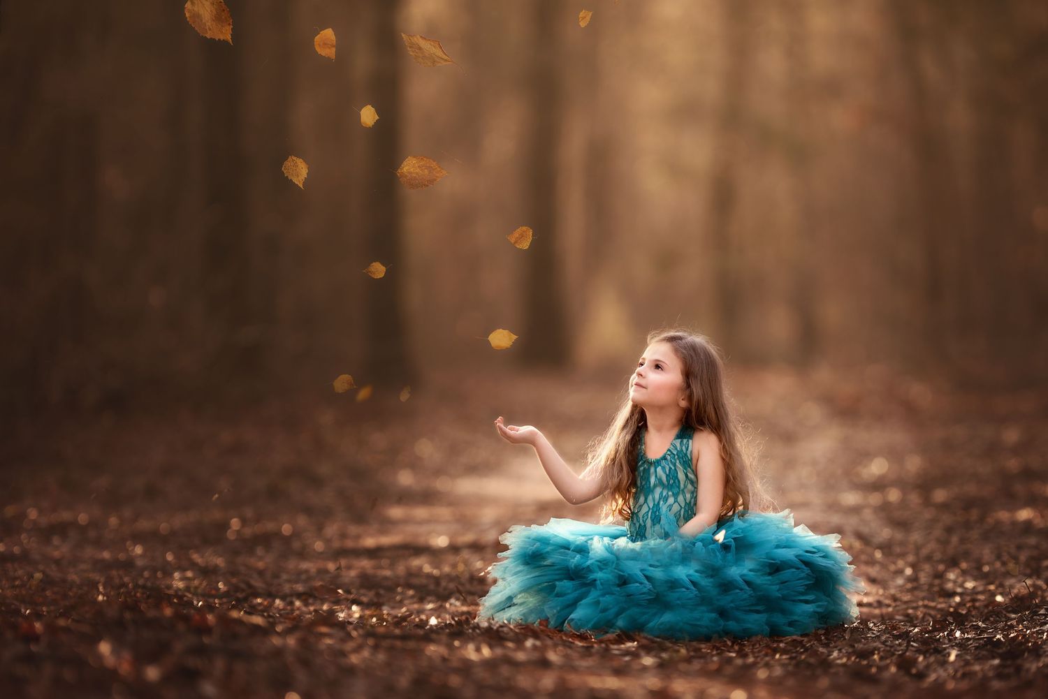 A person in a vibrant teal dress lies dreamily among autumn leaves in a forest setting as golden sunlight filters through.